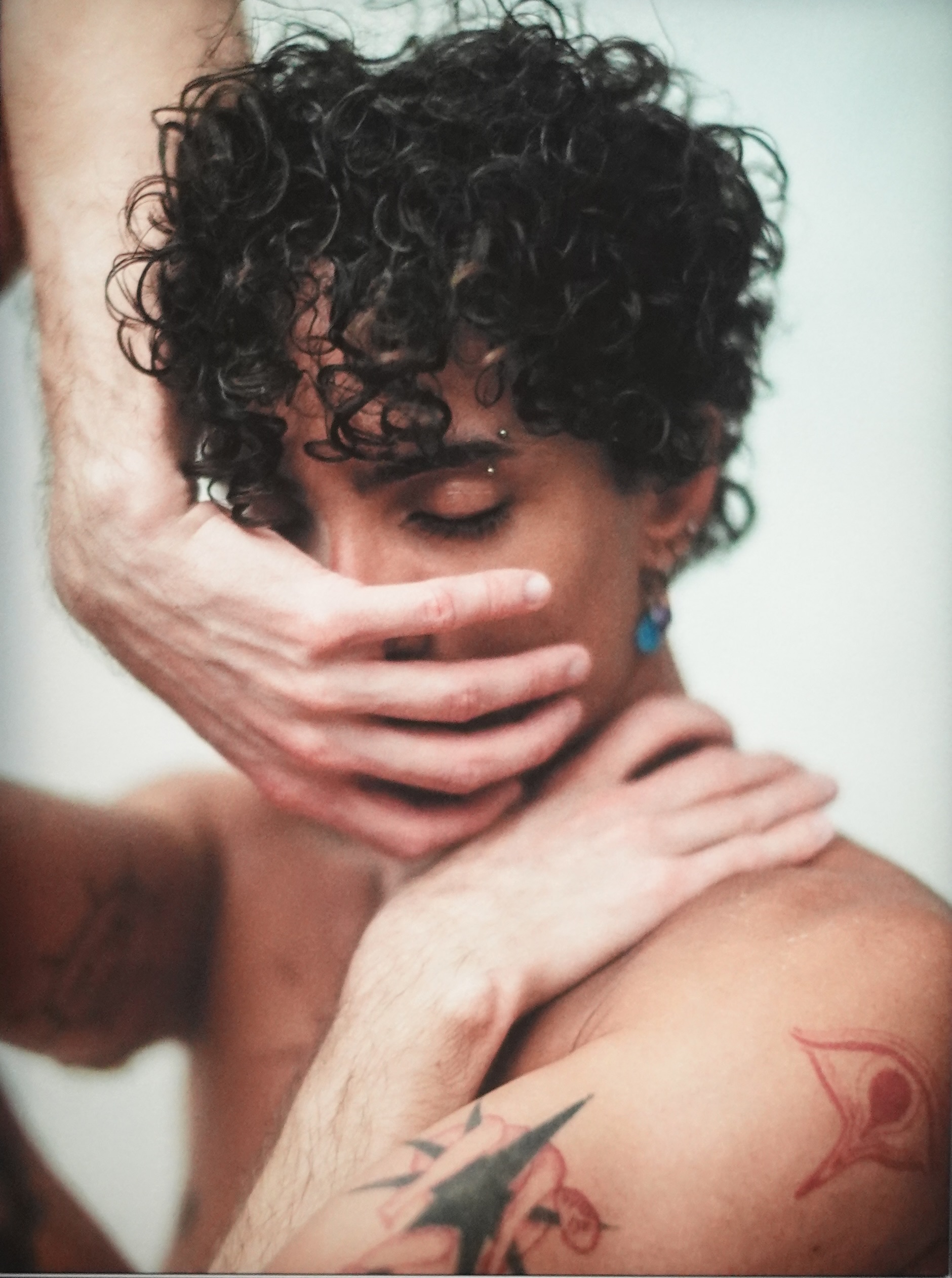 Person mit lockigem schwarzem Haar, von hinten fotografiert, Gesicht verborgen, trägt blaue Ohrringe und hat Tattoos am Arm.