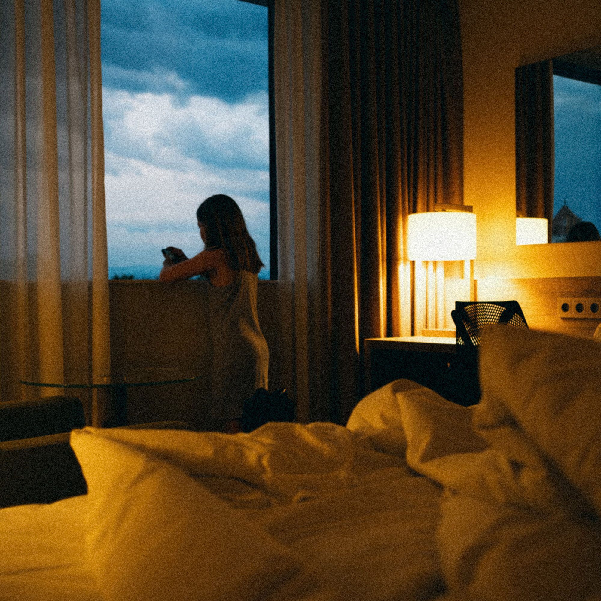 Frau sitzt am Fenster eines Hotelzimmers bei Dämmerung mit Ausblick auf bewölkten Himmel und Landschaft.