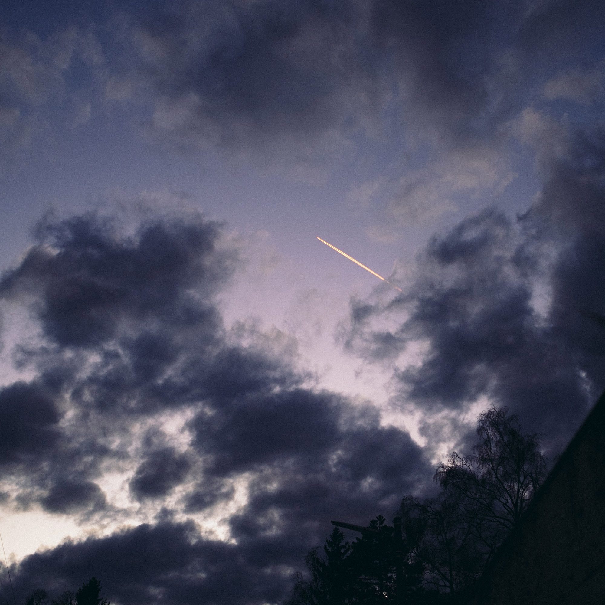Dramatischer Himmel mit dunklen Wolken und einer Lichtlinie (Flugzeug oder Meteor) in der Mitte, Bäume unten.